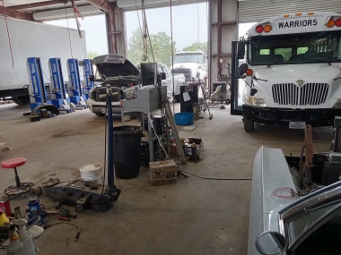 A white diesel bus is facing forward in the Pete's Tire & Automotive Service service garage. The bus has the word Warriors in black at the top. A medium-duty truck is in the background on the left-hand side of the image with its hood popped up for diesel service in Waxahachie, TX